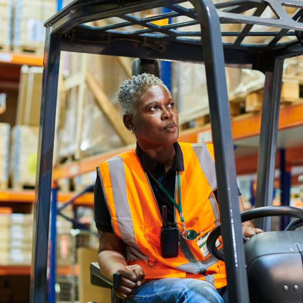 A photograph of a Black female forklift driver with short grey hair wearing a hi-vis vest while driving a forklift truck between tall shelving units